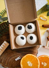 Load image into Gallery viewer, Four bath bombs with black specks in a cardboard box, surrounded by oranges and cinnamon sticks on a decorative surface.