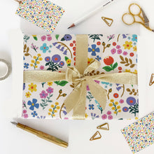 Load image into Gallery viewer, Gift wrapped in floral paper with a gold ribbon on a white surface with stationery items.