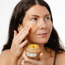 Load image into Gallery viewer, Woman applying balm to her face with a jar of balm in hand on a white background