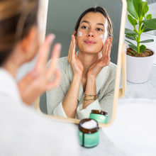 Load image into Gallery viewer, Woman applying cream to her face in front of a mirror with skincare products on a table.