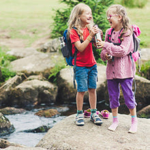Load image into Gallery viewer, Two children with backpacks standing on a rock by a stream, holding bottles.