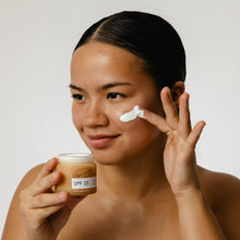 Load image into Gallery viewer, Woman applying sunscreen cream to her face with a jar of sunscreen in her other hand.