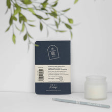 Load image into Gallery viewer, A closed navy blue VVN1 'for change' pocket notebook stands on a white surface with a cream label showing product details. A small frosted candle and a gray pen sit beside it, with soft green leaves hanging from above against a clean white background.