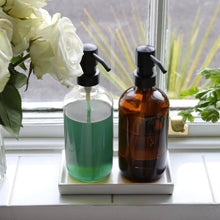 Load image into Gallery viewer, Two bottles with pump dispensers on a windowsill with flowers and plants in the background.
