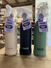 Load image into Gallery viewer, Three Neat refillable cleaning spray bottles standing on a wooden table in a store setting. The bottles are shown in three colors: off-white, dark navy blue, and sage green, each with a white spray top and a purple paper tag.