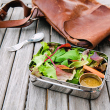 Load image into Gallery viewer, Salad in a metal container with a brown leather bag on a wooden surface