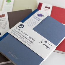 Load image into Gallery viewer, A close-up of a blue and white notebook with a paper band reading 'NOTES, LISTS & SCRIBBLES' and 'VENT for change,' with other notebooks partially visible in the background.