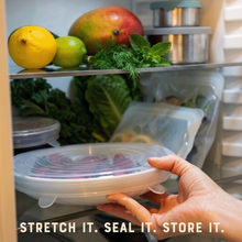 Load image into Gallery viewer, Hand holding a silicone lid over a shelf with fruits and vegetables, with text 'Stretch it. Seal it. Store it.'