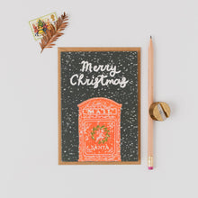 Load image into Gallery viewer, A Christmas greeting card with a dark green snowy background, white 'Merry Christmas' script, and an orange Santa mail mailbox illustration. The card is styled in a flat lay with a brown pencil, a gold pencil sharpener, and a small decorative pine branch on a light gray background.