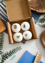 Load image into Gallery viewer, Four white bath bombs with greenery in a brown box on a decorative surface with plants and books.
