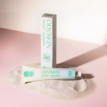 Load image into Gallery viewer, OdySkin sunscreen tube and box on a sand-like surface with a soft pink background
