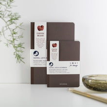 Load image into Gallery viewer, Two brown notebooks with white labels on a light surface with a plant in the background
