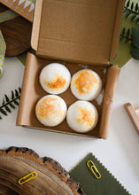 Load image into Gallery viewer, Four bath bombs with orange specks in a cardboard box on a white surface with greenery.
