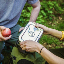 Load image into Gallery viewer, Two people holding a metal lunch box with a logo in a forest setting.
