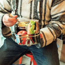 Load image into Gallery viewer, Person holding a metal lunch box with food, sitting on a bench.
