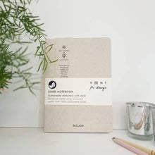 Load image into Gallery viewer, A beige Reclaim lined notebook with a white paper band label, styled on a white surface with a green plant leaf on the left and a glass candle holder on the right.
