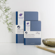 Load image into Gallery viewer, Two blue notebooks with lavender flower branding on a white surface with a plant and candle in the background.
