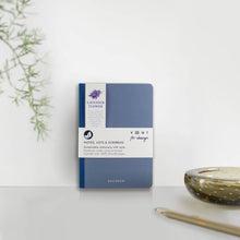 Load image into Gallery viewer, Blue notebook with white label on a light gray surface with a plant and candle in the background
