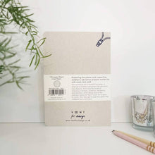 Load image into Gallery viewer, The back cover of a Vent for Change 135 pages Ream sized lined paper notebook, displayed on a white surface next to a glass candle holder and pencils, with green leaves on the left.
