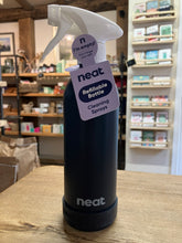 Load image into Gallery viewer, A matte black Neat refillable cleaning spray bottle with a white trigger sprayer and a purple hangtag, displayed on a wooden table inside a gift shop.
