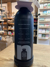 Load image into Gallery viewer, A matte black refillable spray bottle with a lavender spray nozzle and base, standing on a wooden table inside a store. The back of the bottle features white text and a gray graphic with a QR code.
