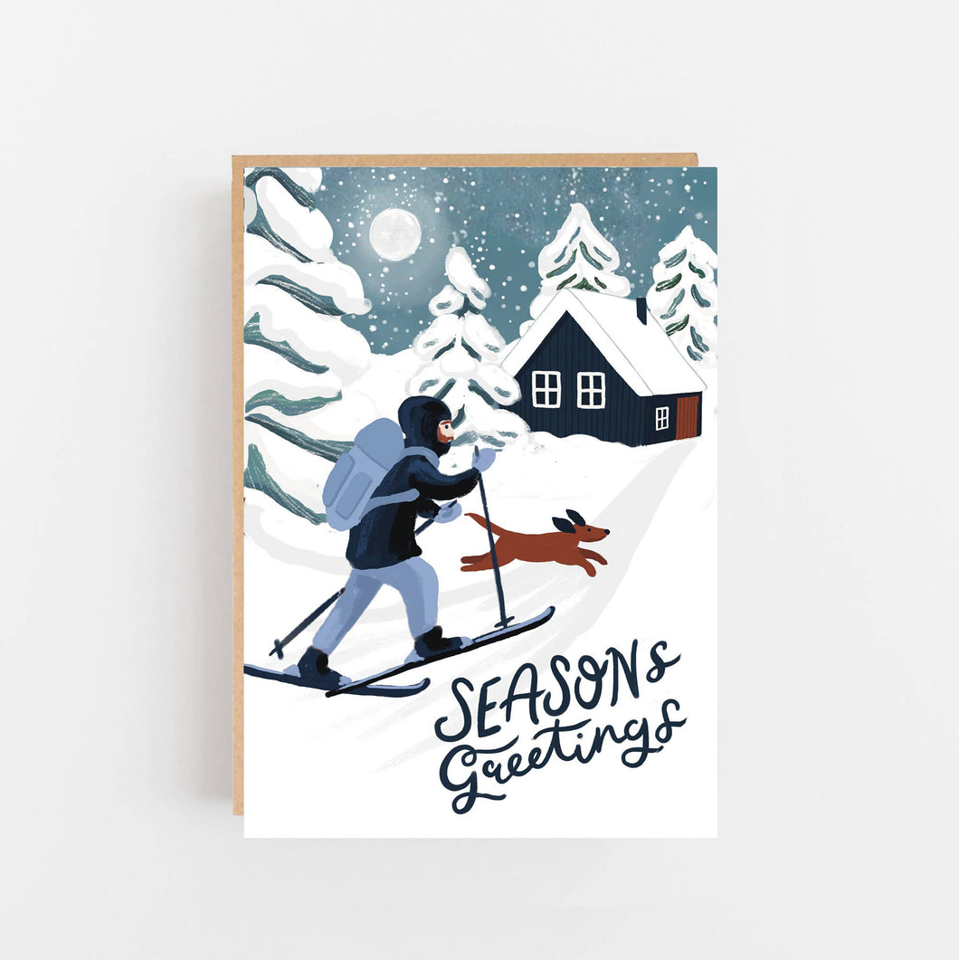 Greeting card with a winter scene of a person skiing with a dog, a house, and trees in the background, with 'Seasons Greetings' text.