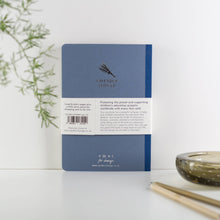 Load image into Gallery viewer, Back cover of a notebook with branding and text on a white surface with plants and a candle.
