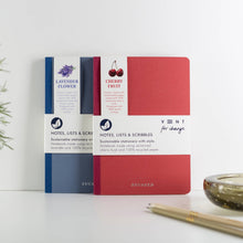 Load image into Gallery viewer, Two VENT for change notebooks, one lavender flower and one cherry fruit, standing upright on a white surface against a white background.
