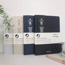 Load image into Gallery viewer, Four Reclaim lined notebooks in different colors (white, beige, navy blue, and dark charcoal) arranged on a white desk with a pencil and plant in the background.
