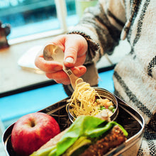 Load image into Gallery viewer, Person eating noodles from a bento box with an apple and corn on a blurred outdoor background
