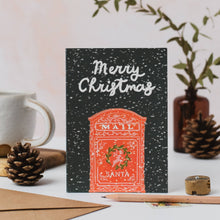 Load image into Gallery viewer, A Christmas greeting card standing upright on a tabletop, featuring the words “Merry Christmas” above an orange-red mail box with Santa and a wreath. The card is styled with pinecones, a mug, a pencil, and a small vase in the background.

